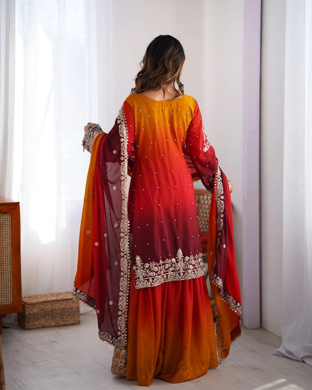 Woman wearing a red and orange traditional outfit with intricate designs in a bright room.