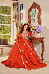 Woman in a red saree with gold details standing in a room with a decorative mirror and floral arrangement.