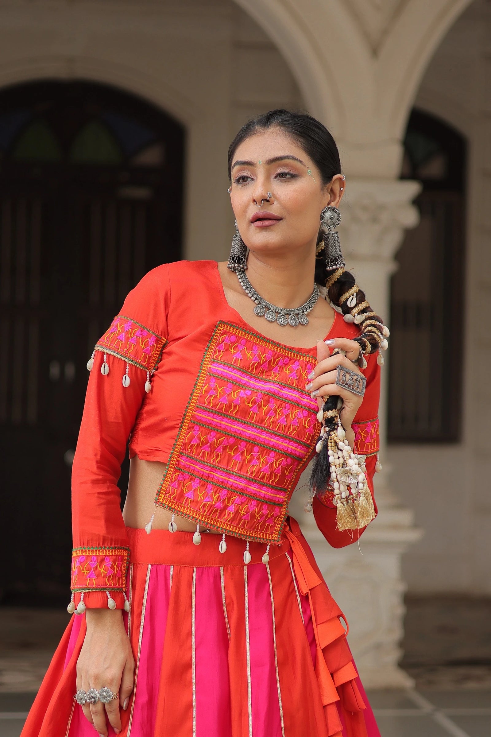Woman in traditional red and pink outfit with jewelry, holding a decorative item, against an architectural background.