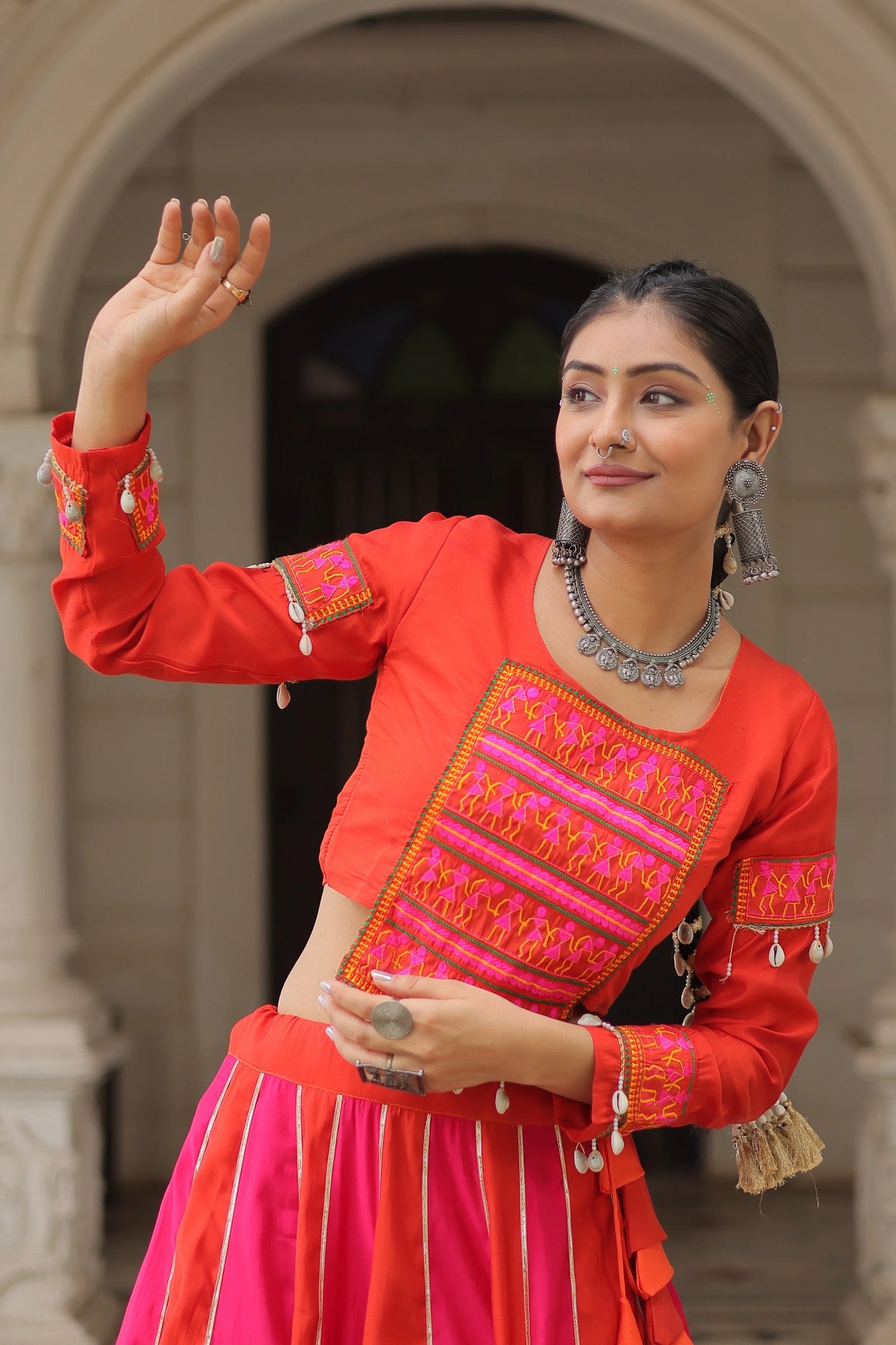 Woman in traditional red and pink outfit with intricate patterns, standing in an archway.