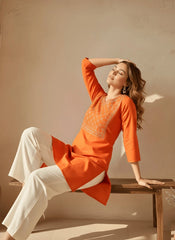 Woman in an orange kurta with white pants sitting on a wooden stool against a beige wall.