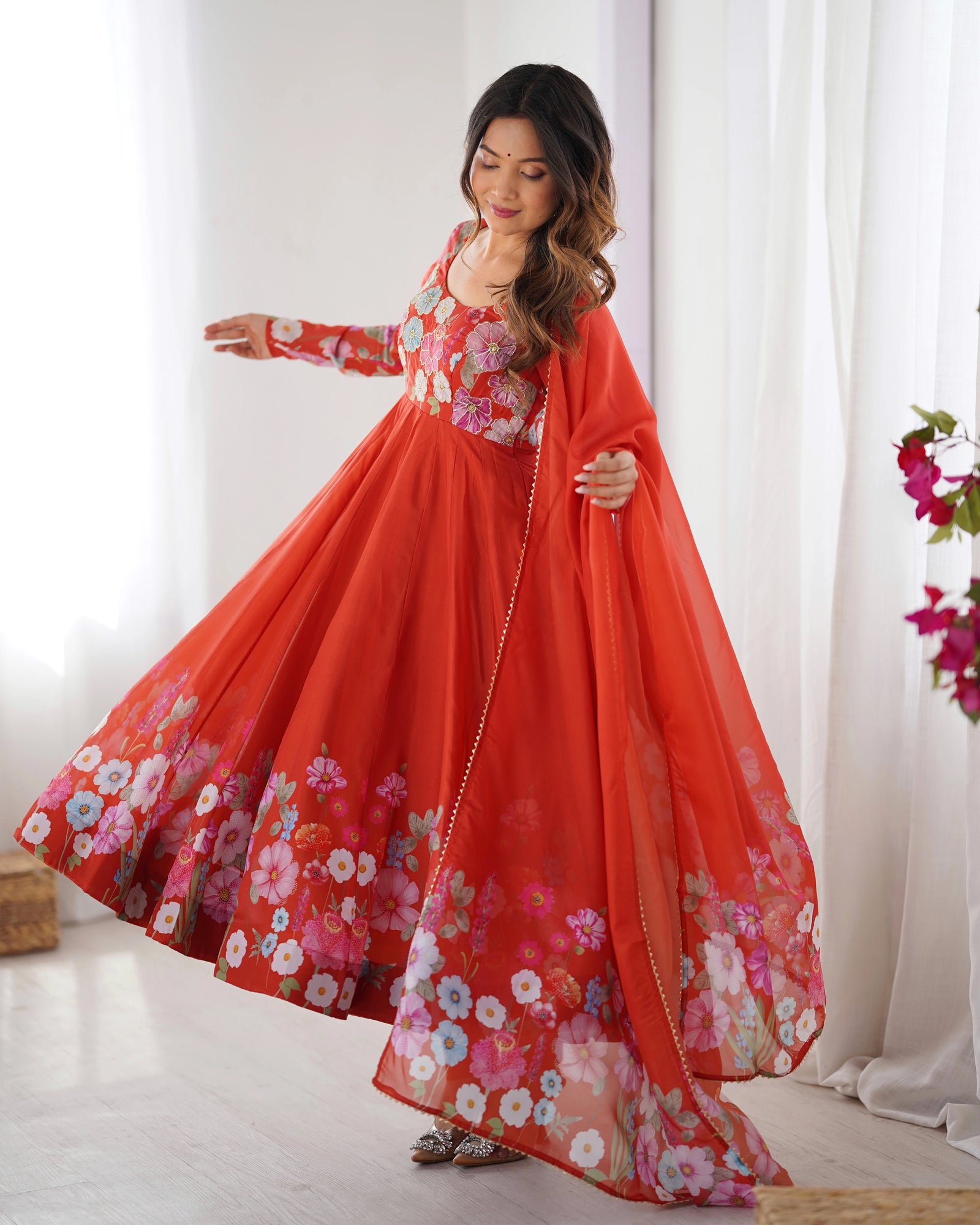 Woman wearing a red floral dress in a bright room with white curtains.