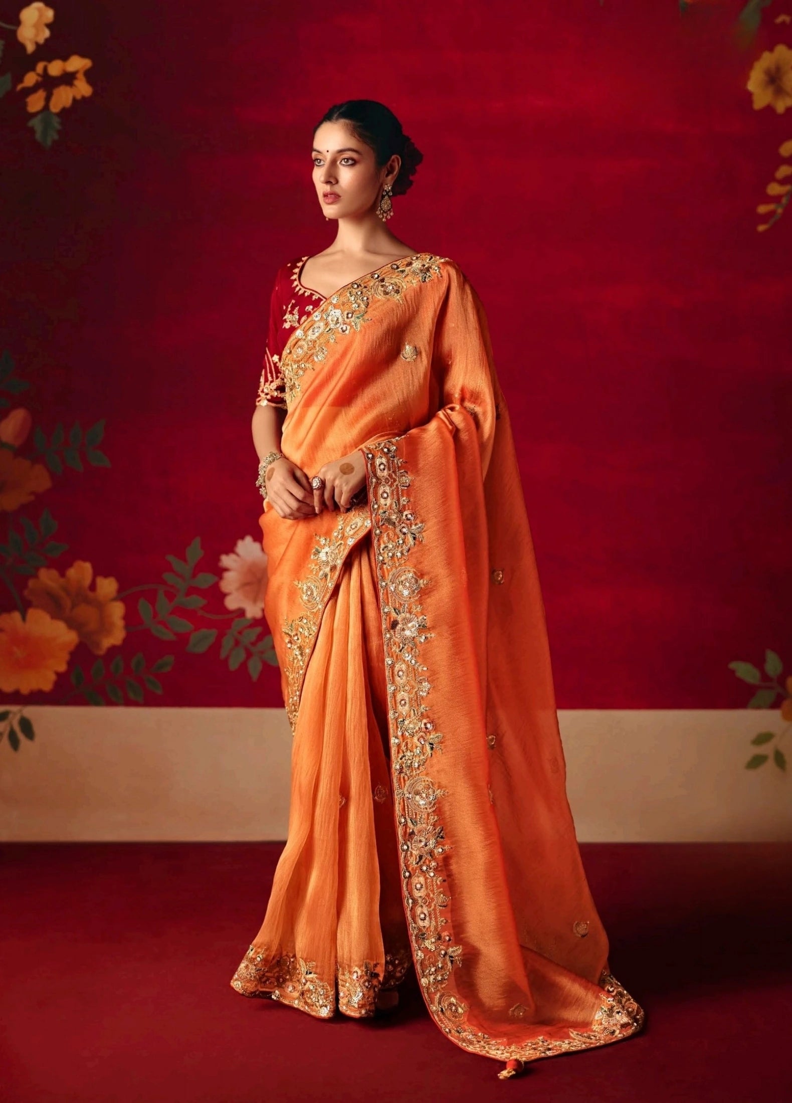 Woman in an orange saree with gold embroidery against a red floral background