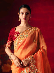 Woman wearing a traditional orange and red saree with gold embroidery against a red background