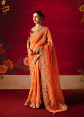 Woman in an orange saree with gold embroidery against a red floral background