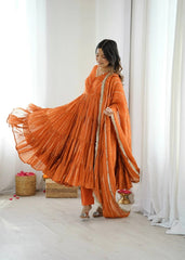Woman in an orange traditional outfit standing in a bright room with white curtains.