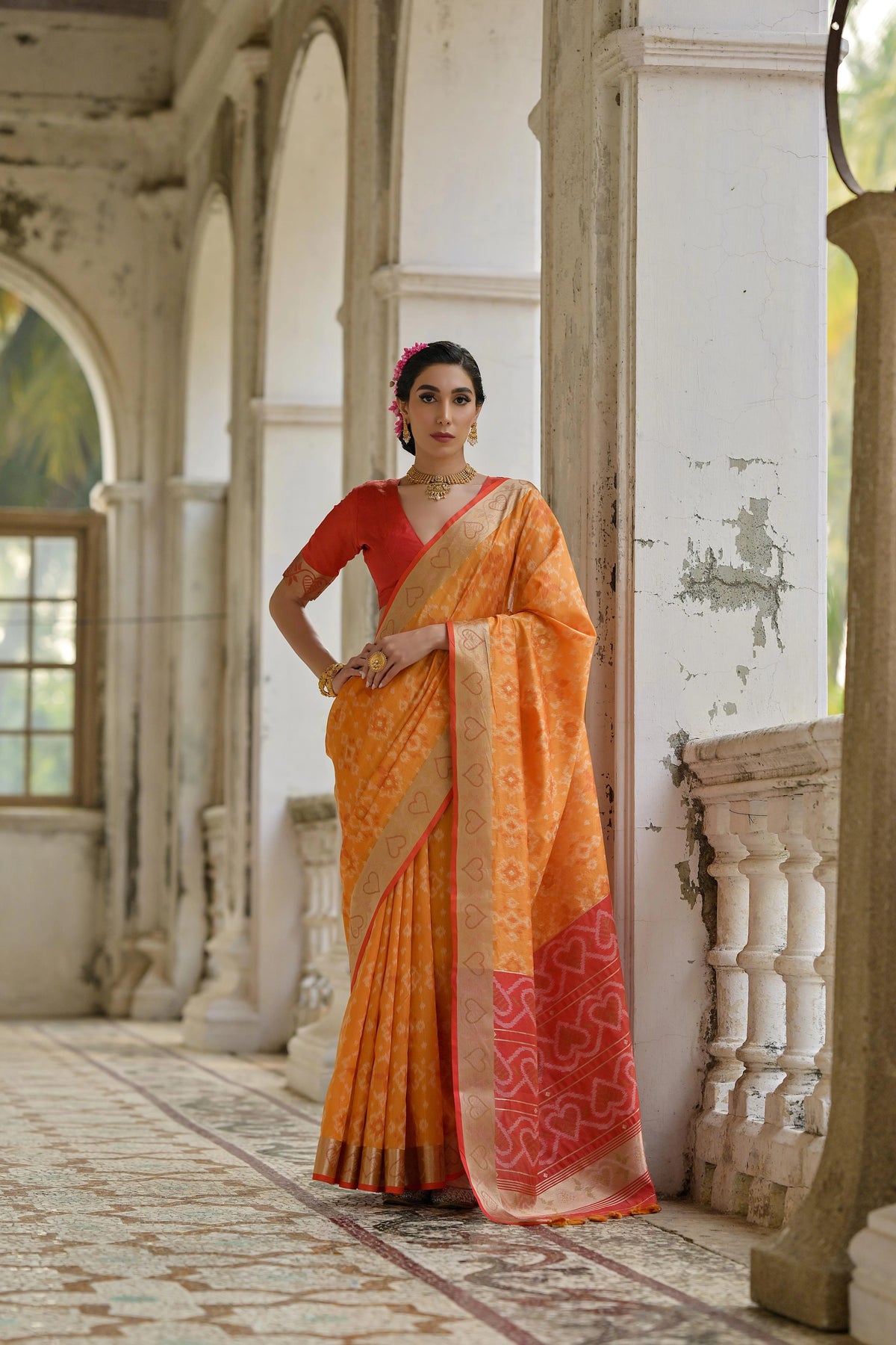 Woman in an orange and red saree standing in a decorative indoor setting.