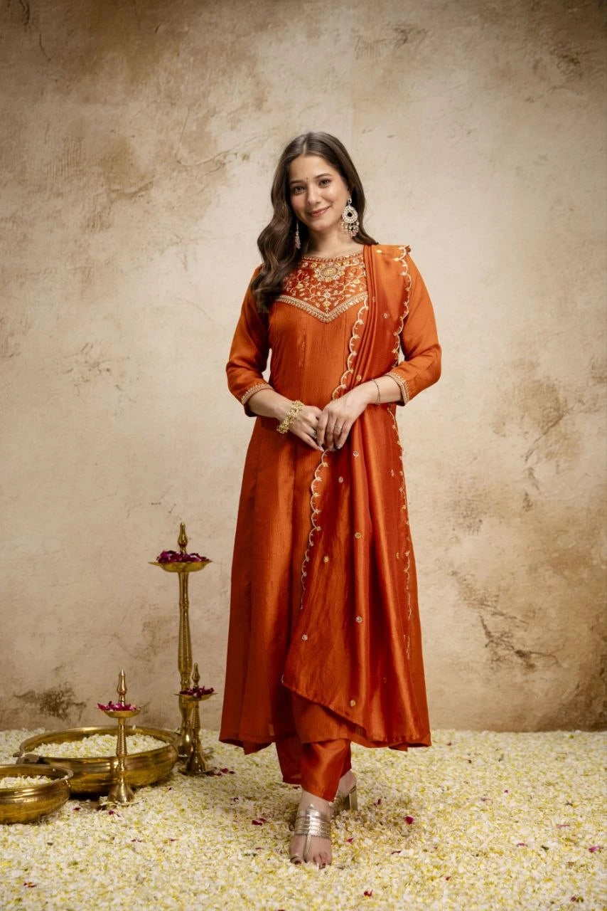 Woman in an orange traditional outfit standing against a textured beige wall with decorative items on the floor.