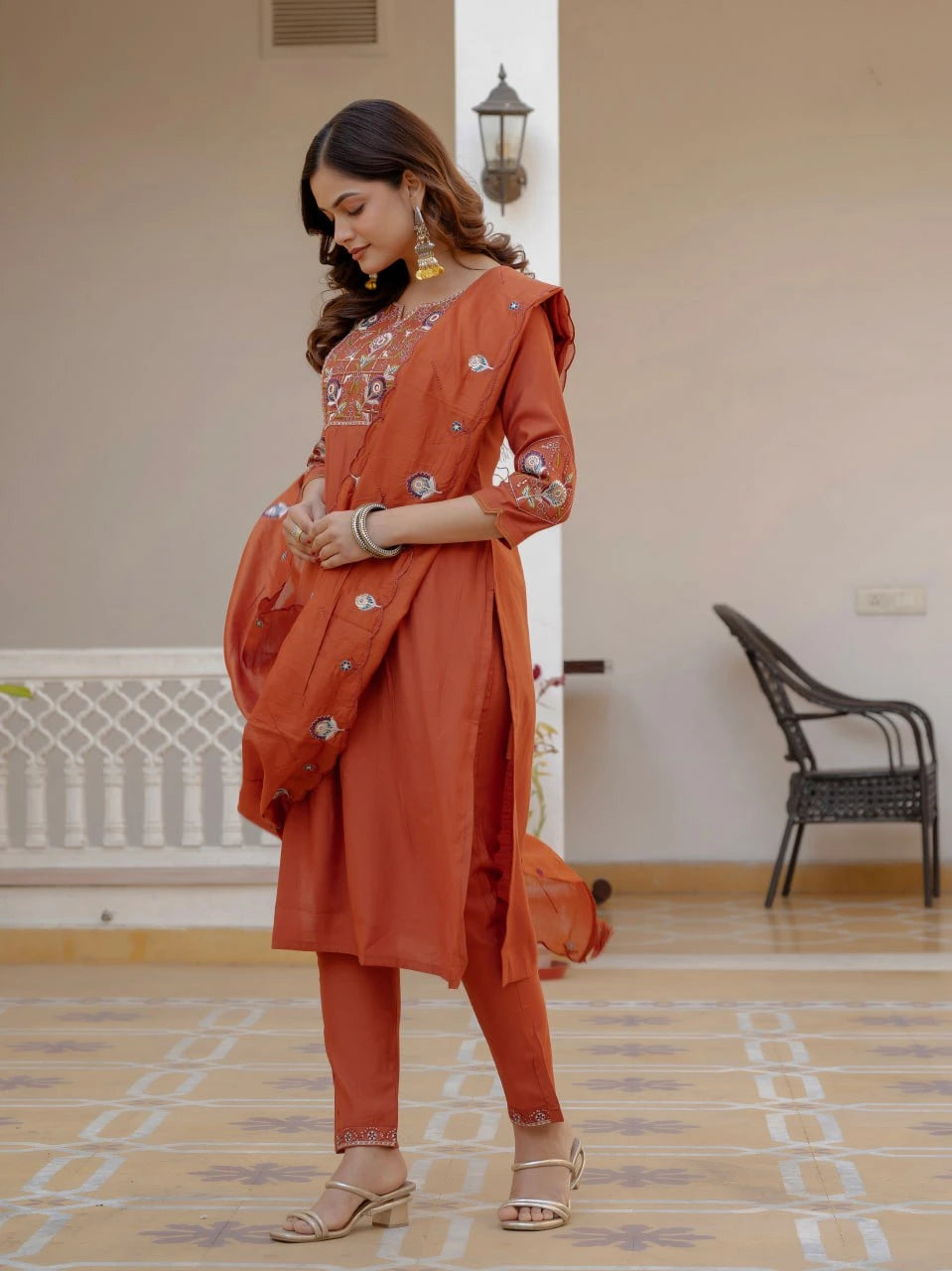 Woman in an orange traditional outfit standing in a room with a chair and lamp in the background.