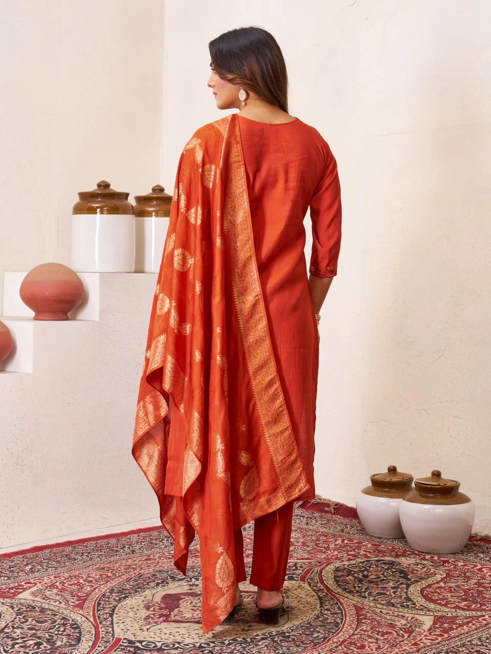 Woman in an orange traditional outfit standing in a room with decorative pots and a patterned rug.