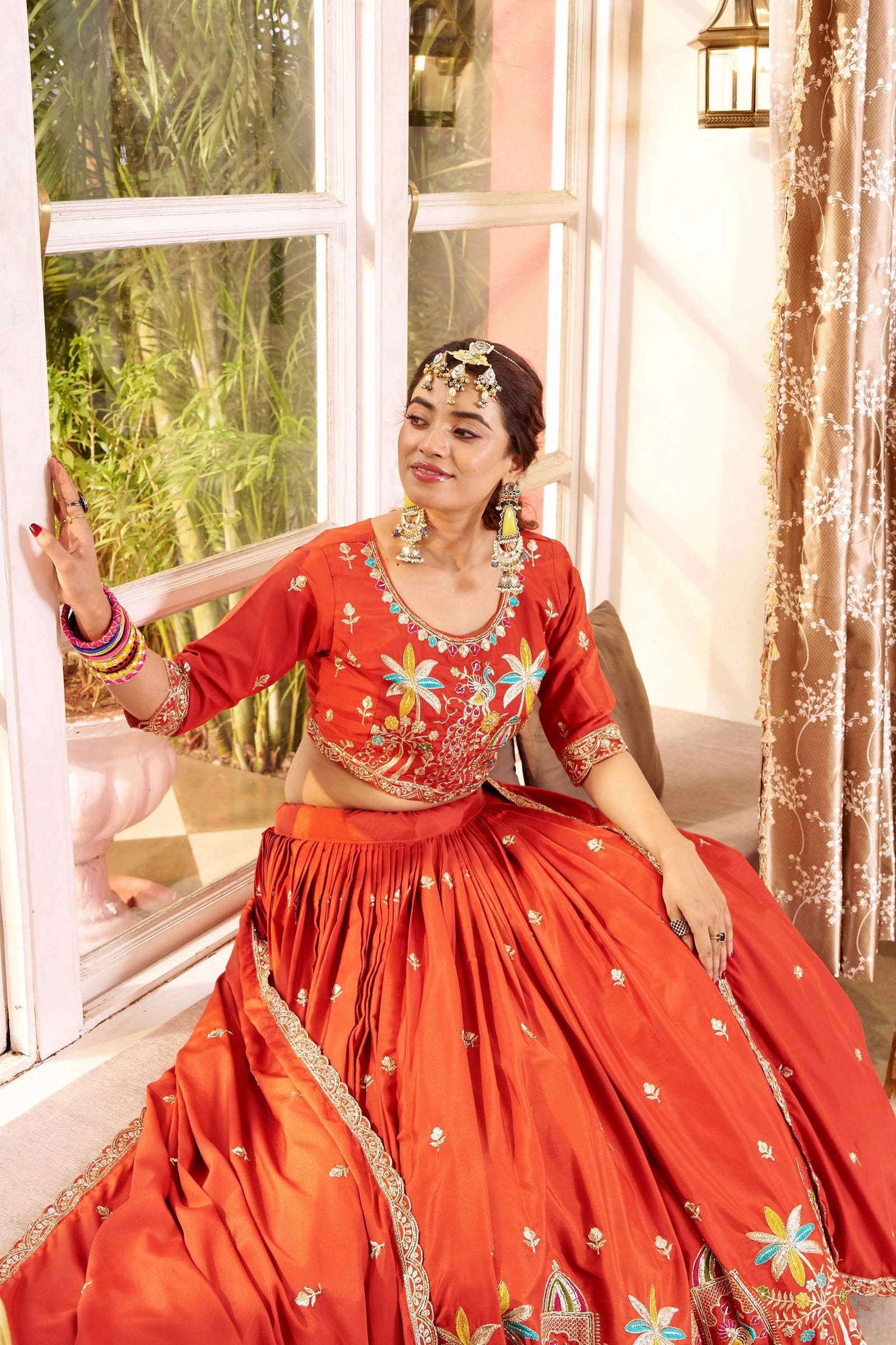 Woman in a red traditional outfit with floral patterns sitting by a window.