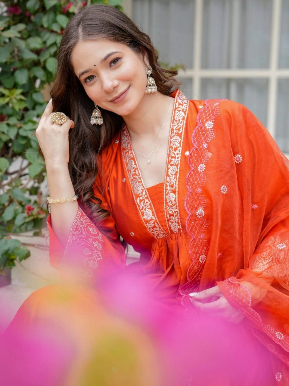 Woman in an orange traditional outfit with floral patterns, sitting outdoors.