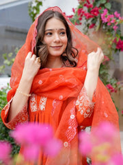Woman wearing a bright orange traditional outfit with floral patterns, standing outdoors with blurred flowers in the foreground.