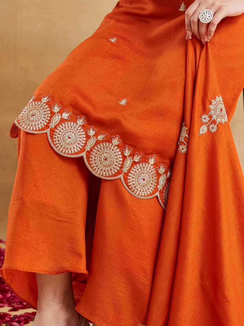 A close-up detail shot of the lower portion of a rust-orange silk kurti set, highlighting the intricate embroidery. The heavy, gold-thread embroidery features traditional circular motifs and patterns along the scalloped hemline of the kurta. A matching dupatta is draped, showing similar gold threadwork. A partial view of a woman's leg wearing a large cocktail ring on her finger is included, along with red rose petals on the floor in the foreground.