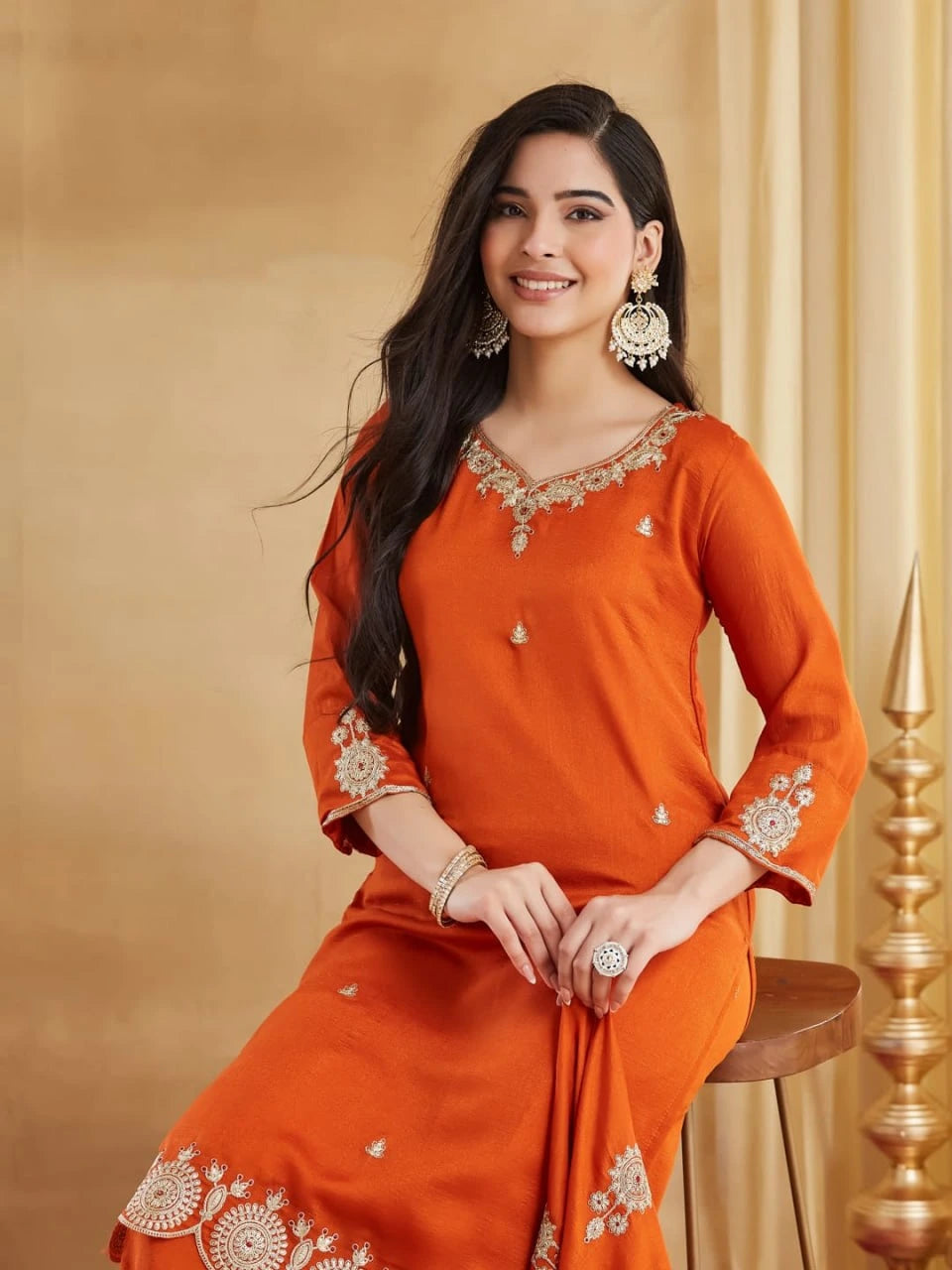 A full-length portrait of a woman seated on a low wooden stool, smiling and facing slightly right. She is wearing an elegant rust-orange kurta and palazzo set. The V-neck kurta is adorned with intricate gold and maroon thread work around the neck and sleeve cuffs. The set features wide embroidered panels at the kurti hem and pants cuffs with traditional motifs. She is adorned with heavy, elaborate gold and pearl jhumka earrings, a stack of bangles, and a large ring.