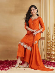 A woman is seated gracefully on a wooden stool, smiling and looking toward the camera. She is wearing a rust-orange Raw Silk Kurti set. The short kurta features intricate gold thread embroidery on the neck, sleeves, and a prominent scalloped embroidered hemline. She has wide-legged trousers and matching juttis. Her look is accessorized with heavy gold traditional earrings, bangles, and a cocktail ring. The floor is scattered with red rose petals, and gold pyramid structures are visible in the background