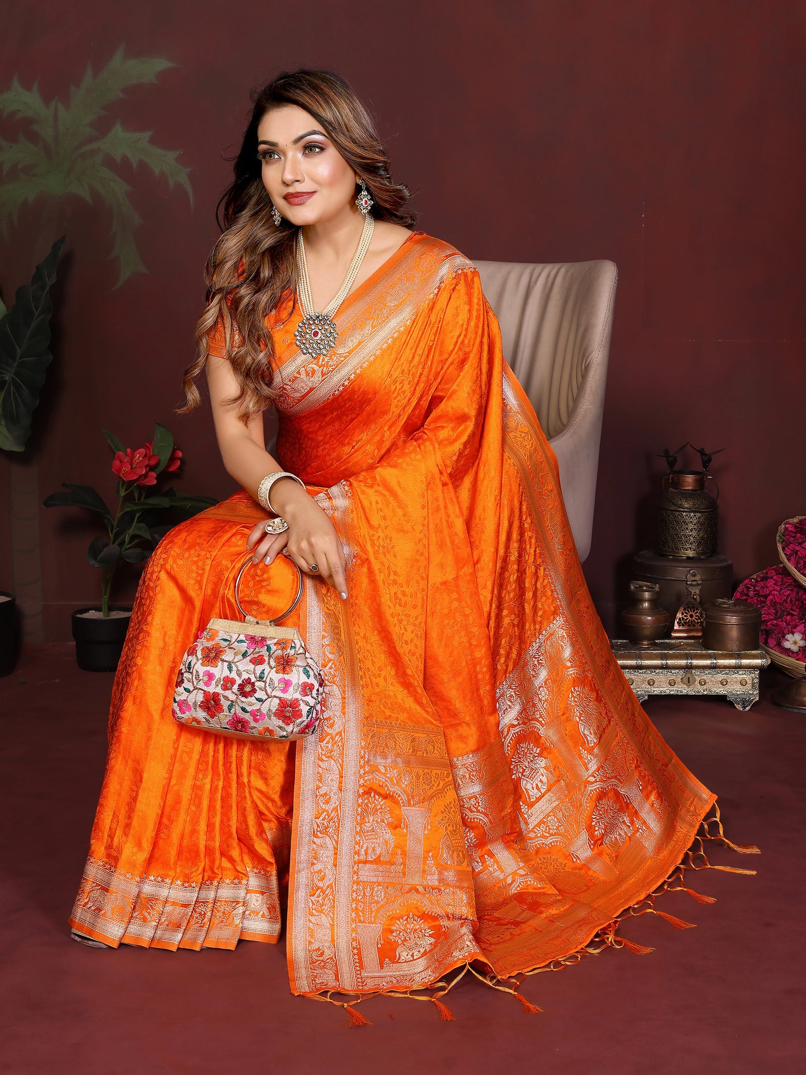 Woman in an orange saree with a decorative bag, sitting against a dark background.