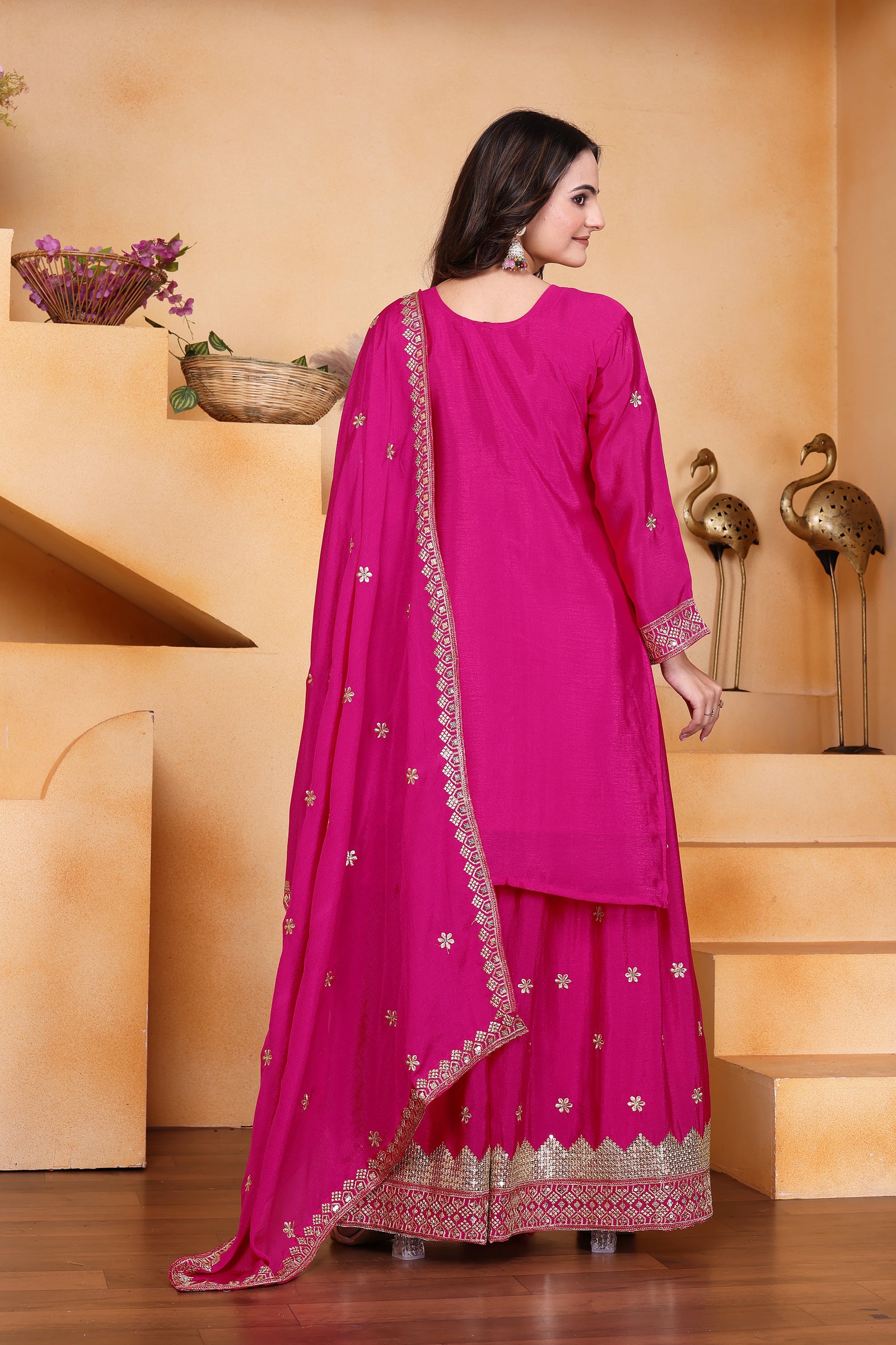Woman in a bright pink traditional outfit with gold details standing in a decorative indoor setting.