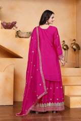 Woman in a bright pink traditional outfit with gold details standing in a decorative indoor setting.