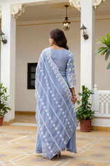 Woman in a blue saree with white patterns walking in an indoor setting with plants and architectural elements.