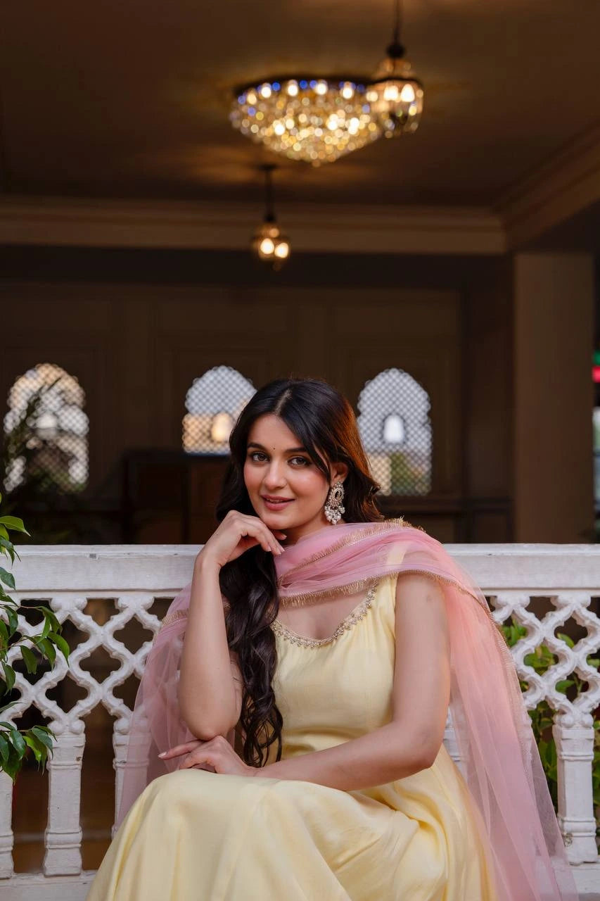 Woman in a yellow dress with a pink dupatta sitting on a white railing indoors.