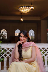 Woman in a yellow dress with a pink dupatta sitting on a white railing indoors.