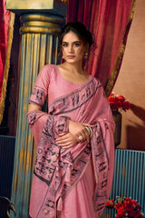 Woman in a pink saree with intricate patterns standing against a decorative background.