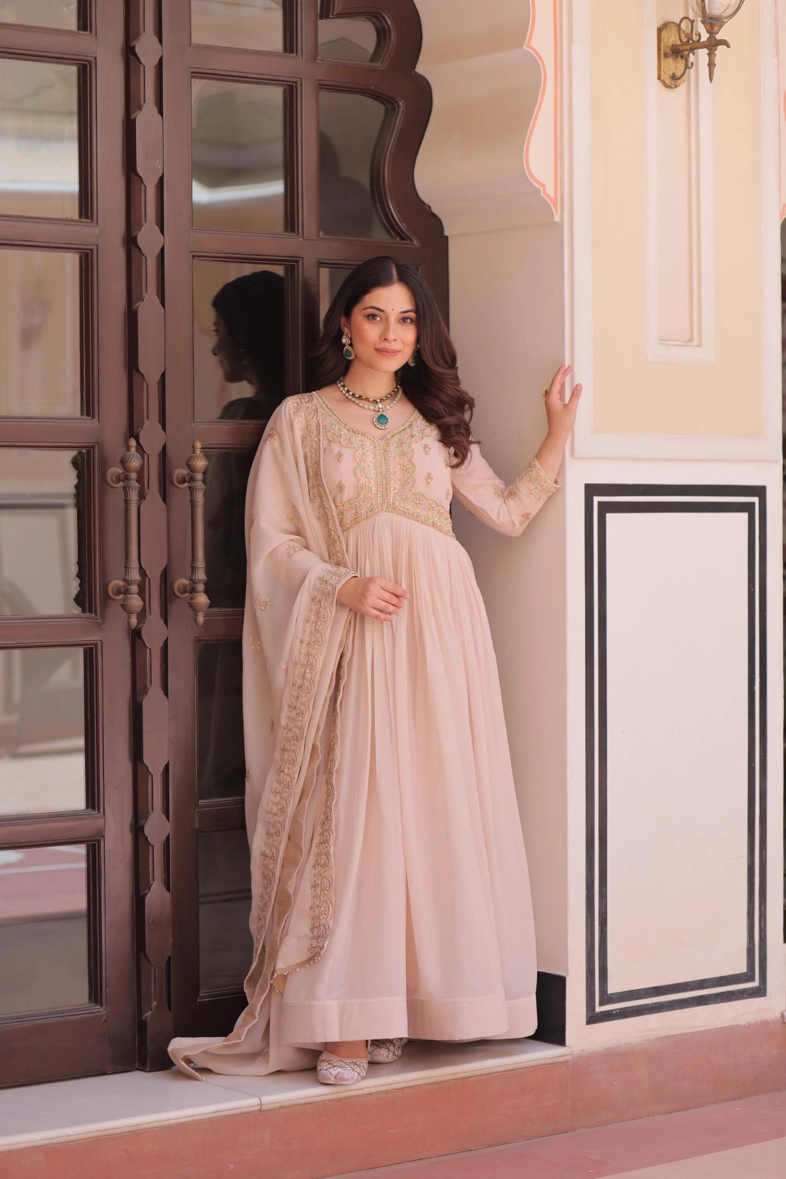 Woman in a beige traditional outfit standing in a doorway with decorative wall and door.