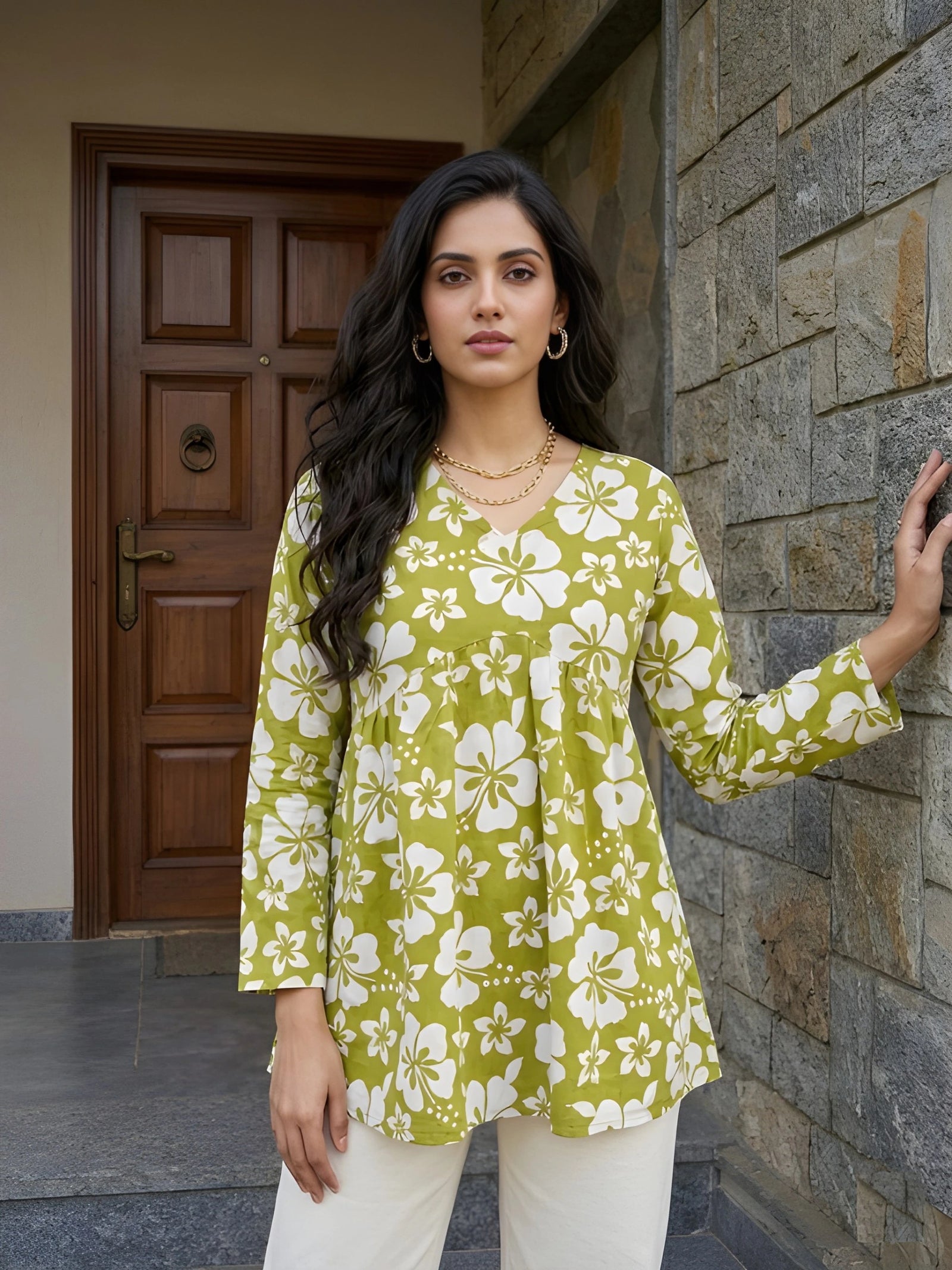 A medium shot of a woman posing in a green floral tunic and white pants, resting one hand against a textured stone wall.