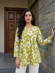 A medium shot of a woman posing in a green floral tunic and white pants, resting one hand against a textured stone wall.