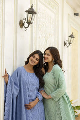 Two women in blue and green lace dresses standing against a white wall with decorative lights.
