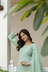 Woman in a light green traditional outfit standing indoors with plants in the background