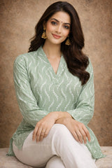 Woman wearing a green patterned top and white pants against a brown background