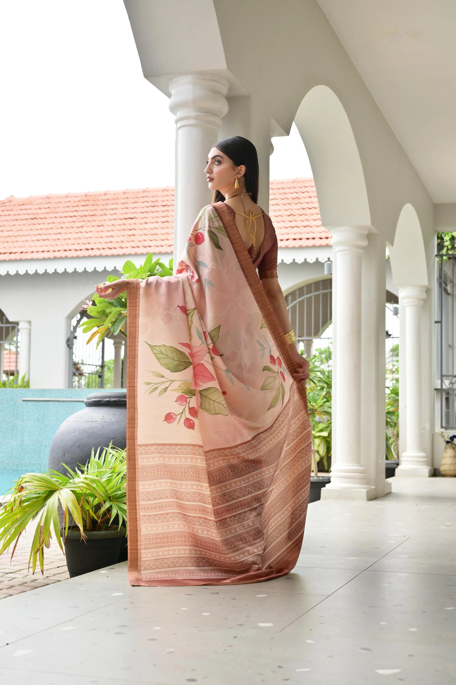 Woman in a floral saree standing on a porch with white columns and greenery.