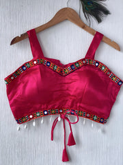 Fuchsia pink ethnic blouse featuring vibrant mirror work and a multicolored embroidered geometric border in green, blue, and orange, adorned with pearl drop tassels and a tie-up waist detail. Displayed on a wooden hanger against a textured off-white wall with peacock feathers, creating a festive and traditional look.