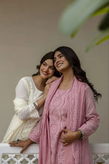 Two women standing close together, one in a white outfit and the other in a pink outfit with lace details, against a neutral background.