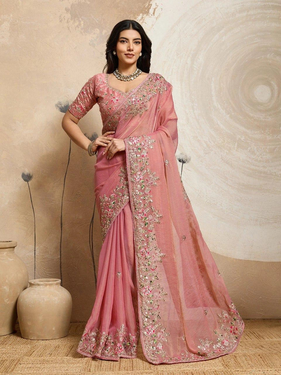 Woman wearing a pink saree with intricate designs against a beige wall.