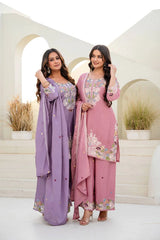 Two women in traditional outfits standing in a decorative setting with plants.