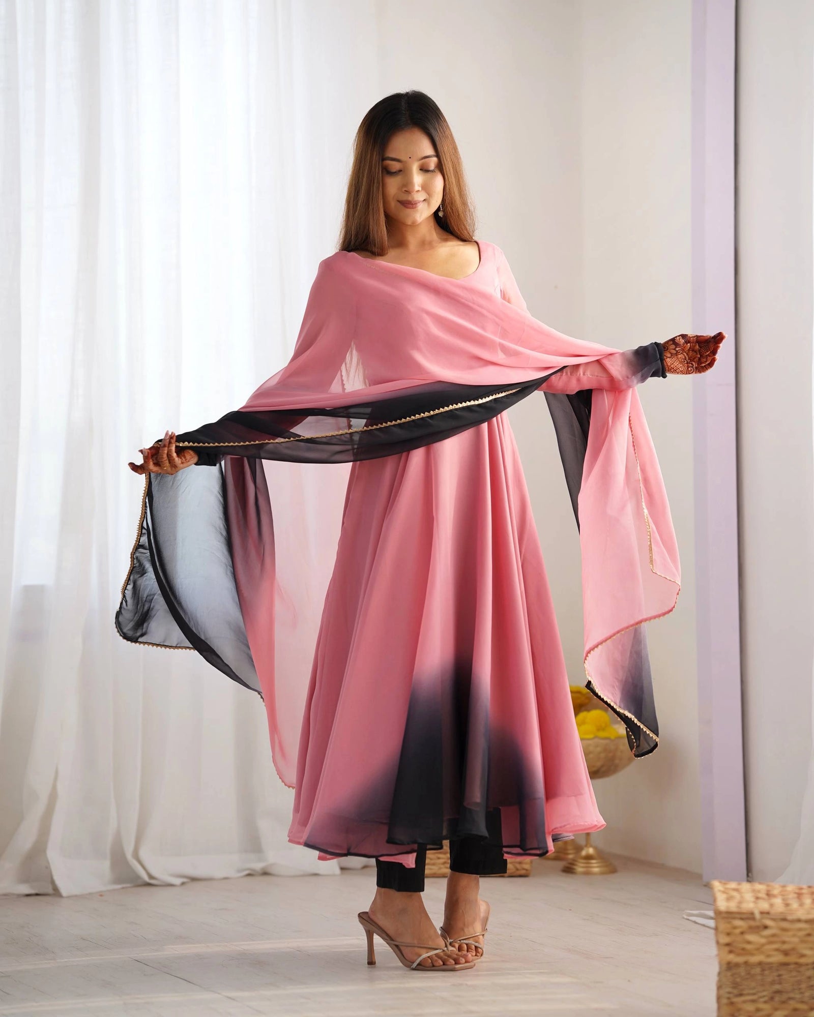 A woman poses standing, looking down while holding a pink and black ombre dupatta draped across her front. Her hands feature intricate henna designs.