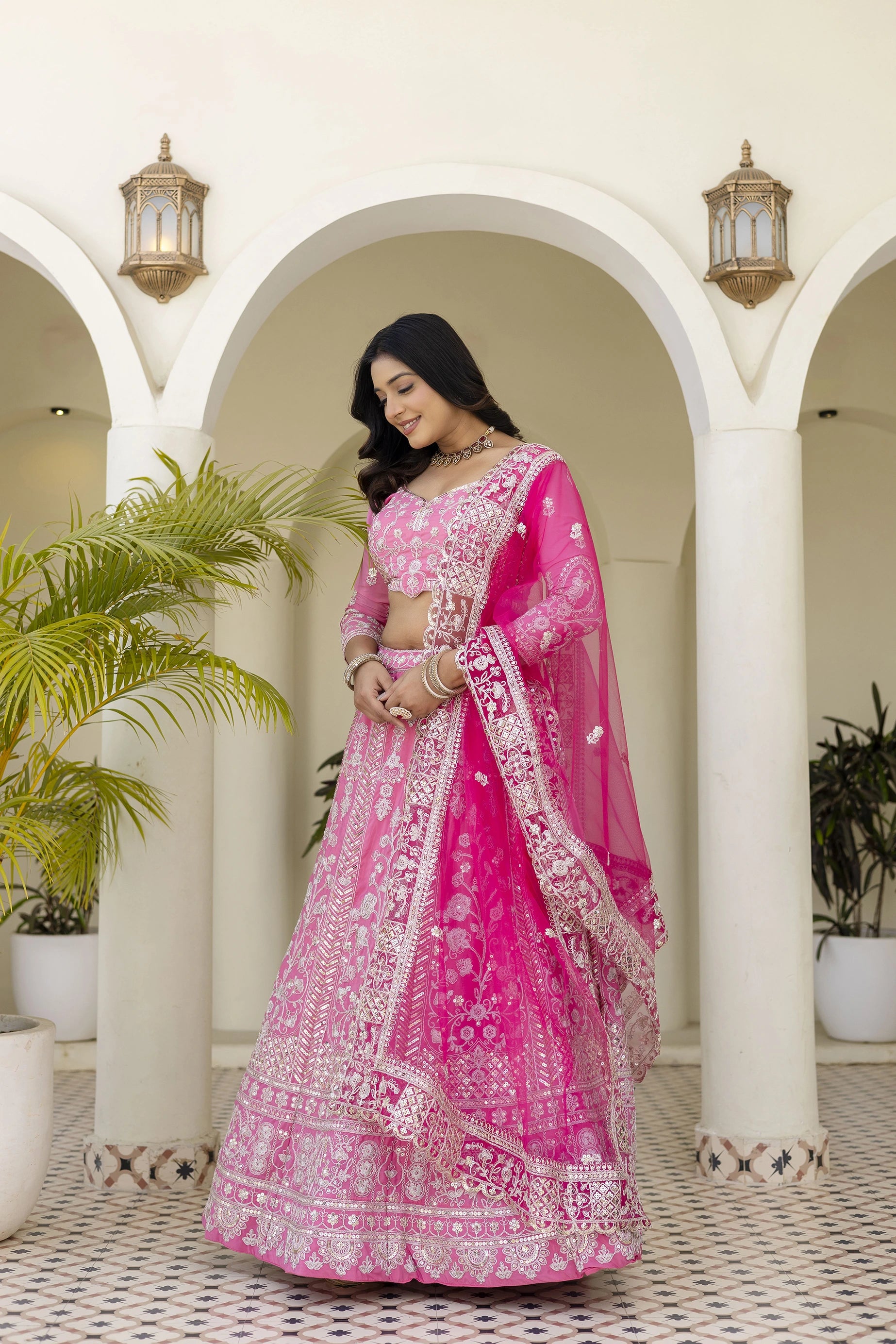 Woman in a pink embroidered traditional outfit standing in an archway with plants.