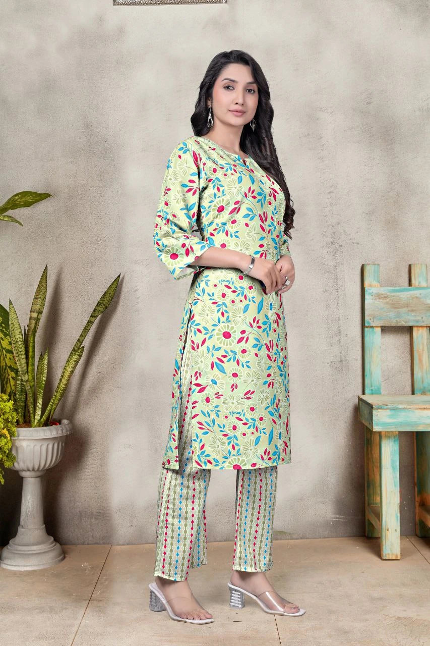 Woman wearing a light green floral outfit standing against a neutral wall with a plant and wooden chair.