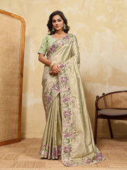 Woman wearing a light green saree with floral patterns in a room with beige walls and a wicker chair.