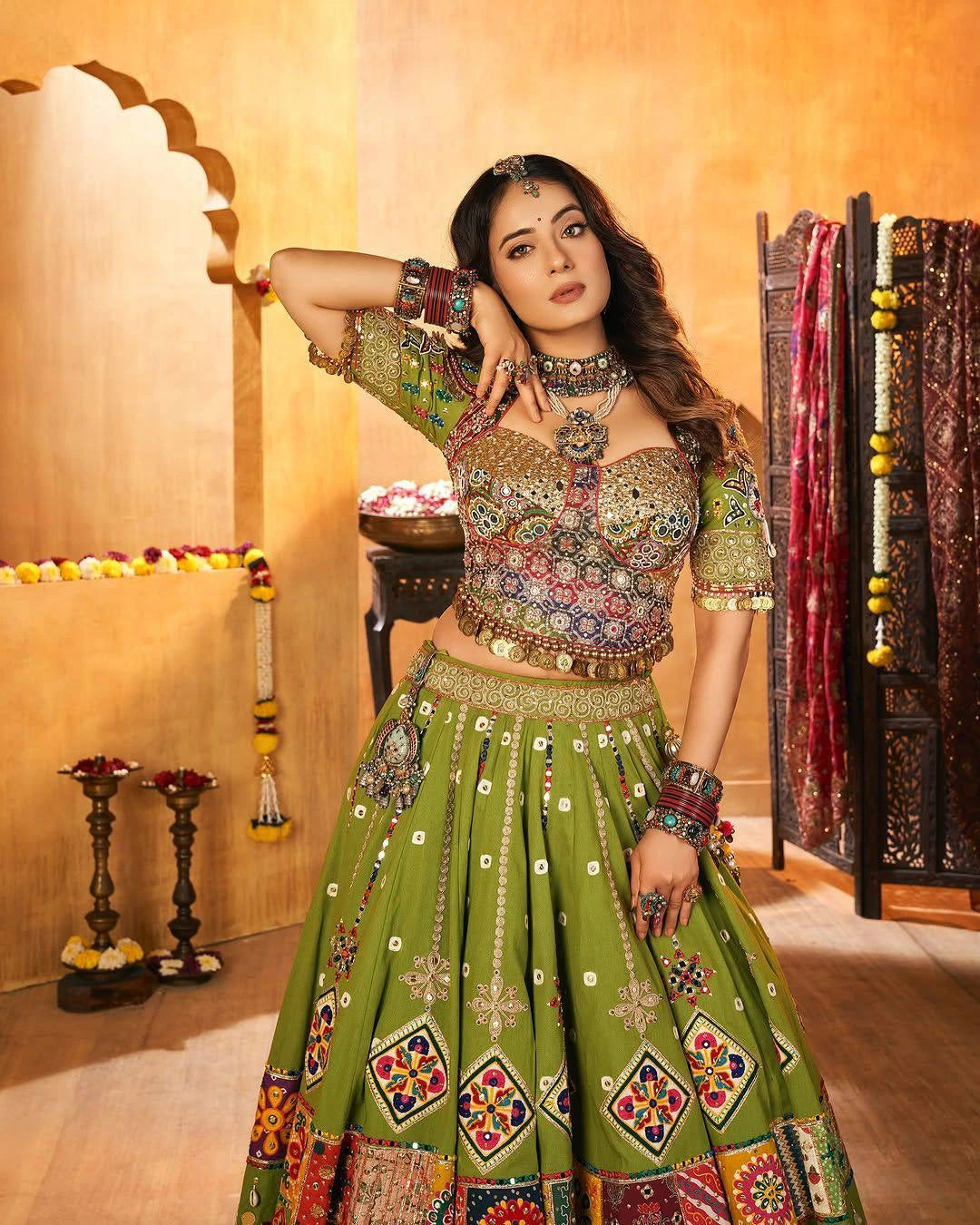 Woman in traditional green and multicolored outfit with intricate patterns in a decorated room.