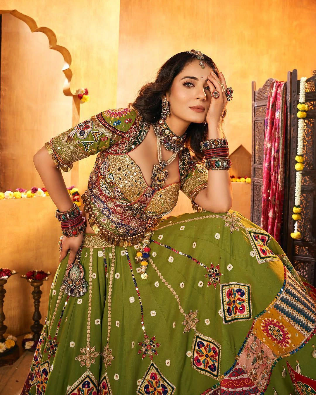 Woman in traditional green and gold embroidered outfit with jewelry in a decorated room.