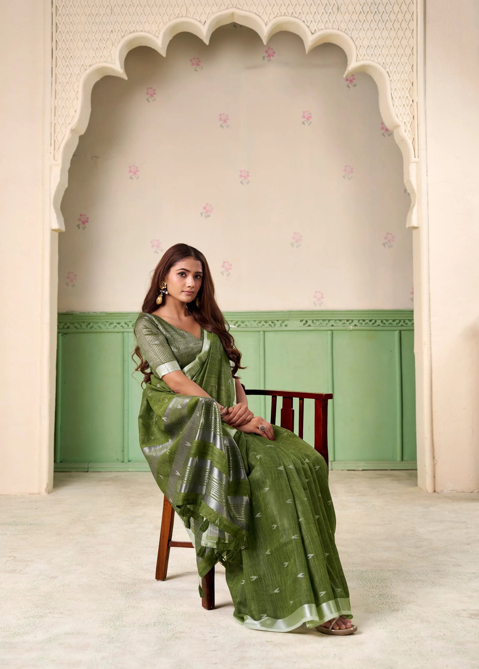 Woman in a green saree sitting in a decorated room with an archway.