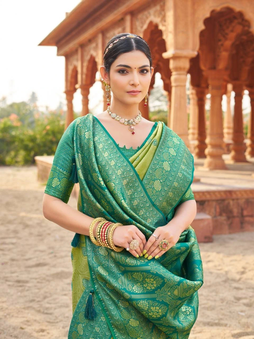 Woman in a green saree with gold patterns standing in front of an architectural structure.