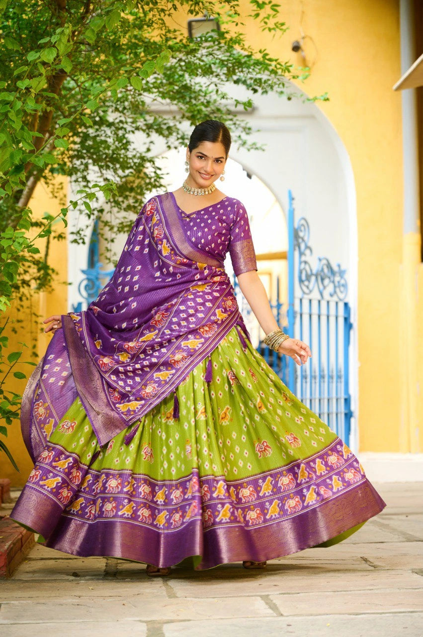 Woman in a green and purple traditional outfit standing outdoors with a yellow wall and greenery in the background.
