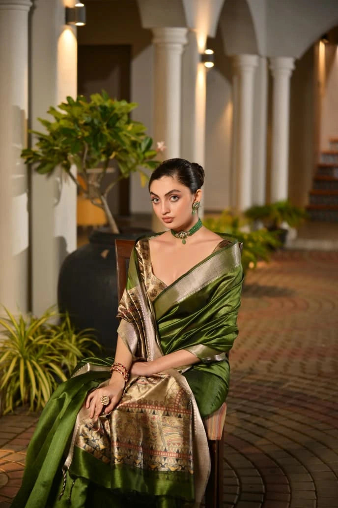 Woman in a green saree sitting outdoors with plants and columns in the background