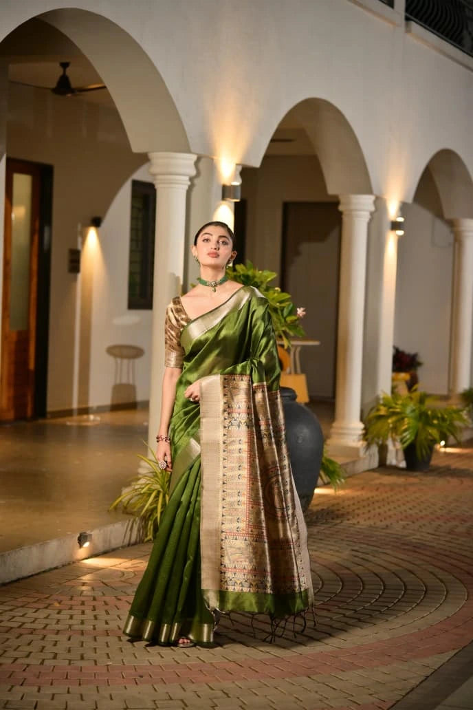 Woman in a green saree standing in an elegant indoor setting with columns and decorative elements.