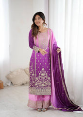 Woman wearing a traditional purple and pink outfit with intricate designs, standing against a white curtain background.